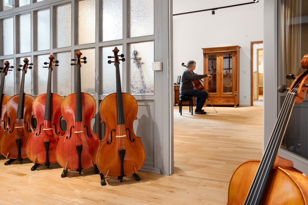 Blick in das Violineo Atelier. Im Vordergrund stehen Celli, im Hintergrund spielt eine Person auf einem Cello.